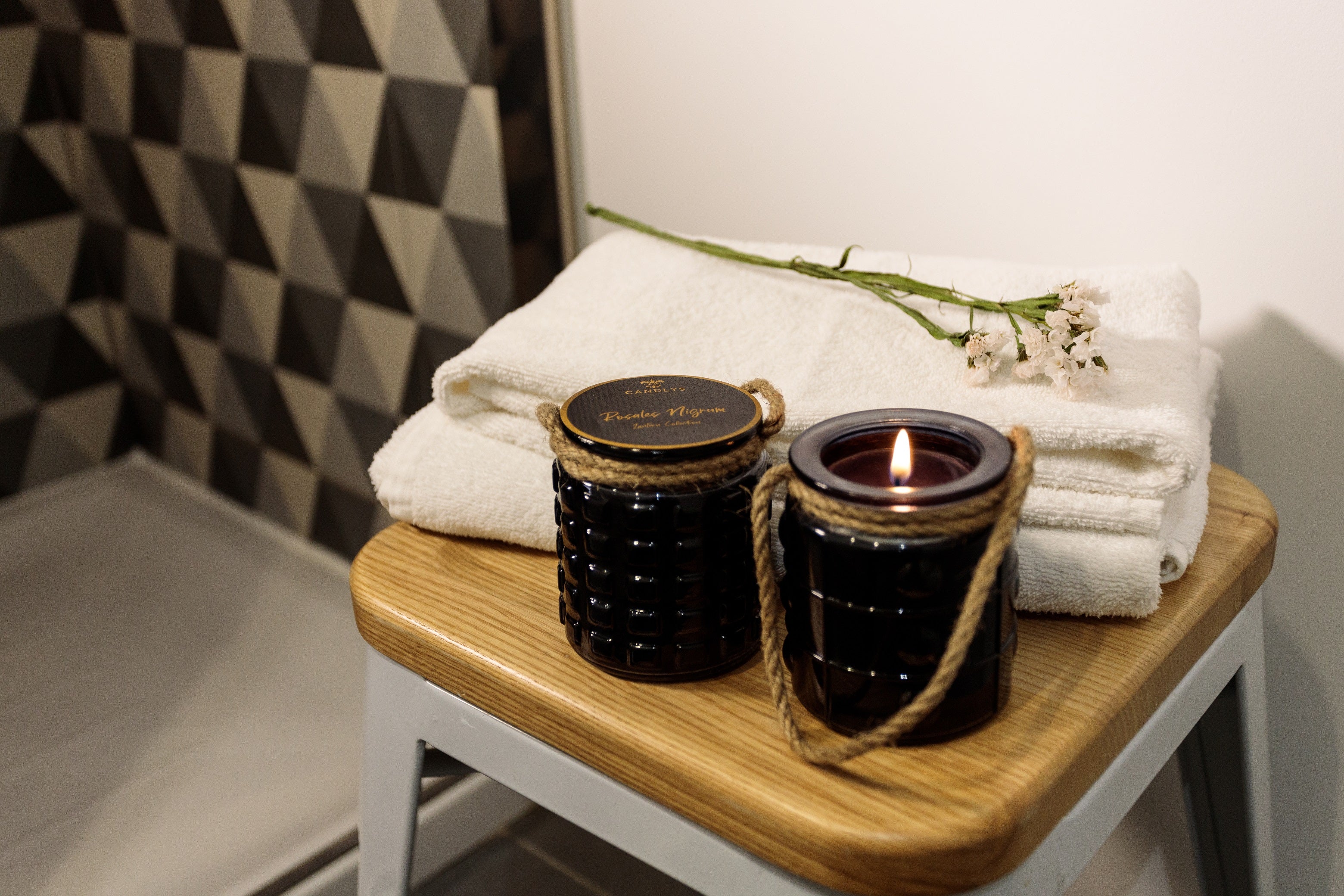Vela aromática preta da Candlys acesa sobre toalhas de spa, com detalhe de flor seca, num ambiente calmo e elegante.