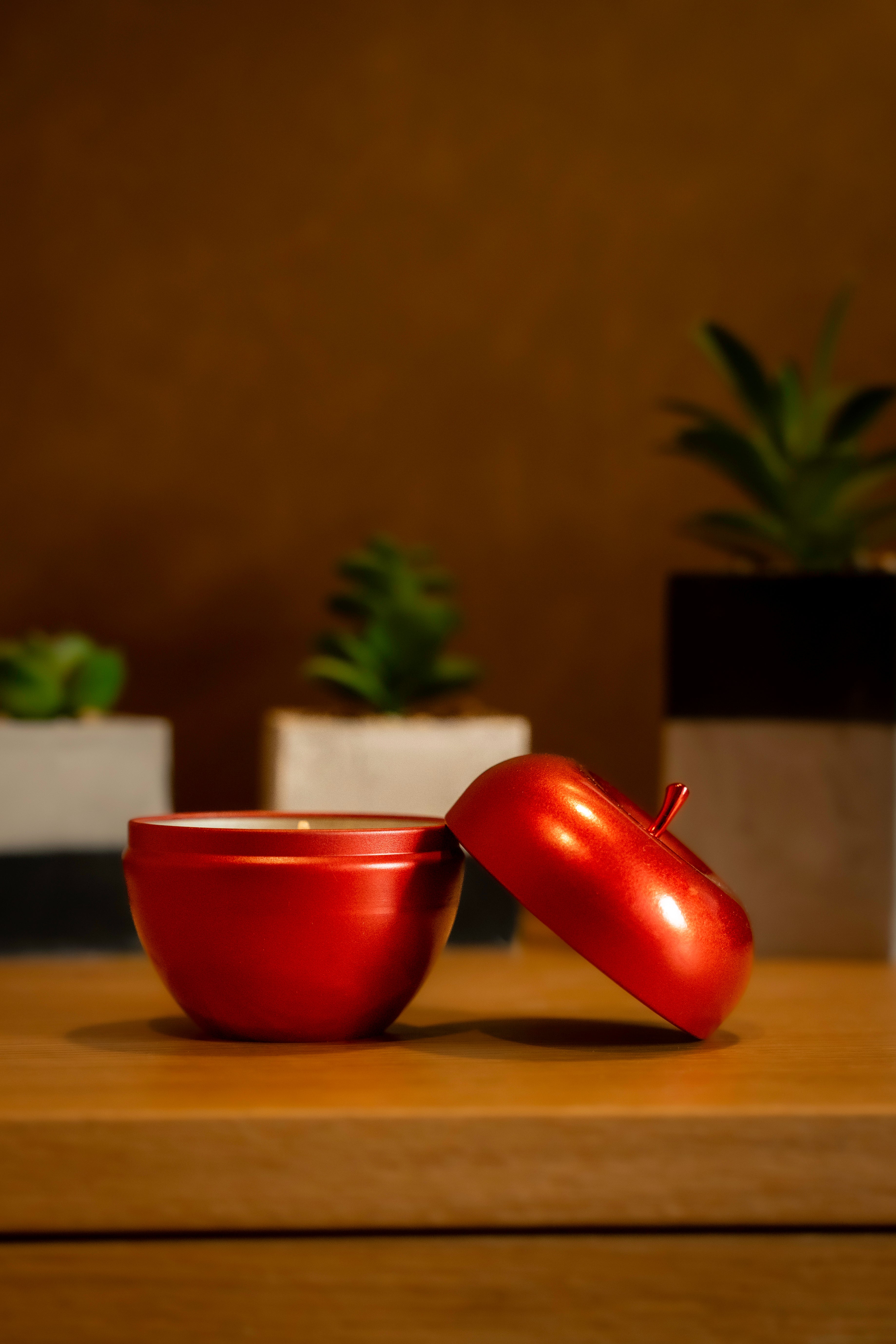 Vela Maçã & Canela em formato de maçã vermelha com tampa aberta, acompanhada por plantas decorativas em fundo aconchegante.