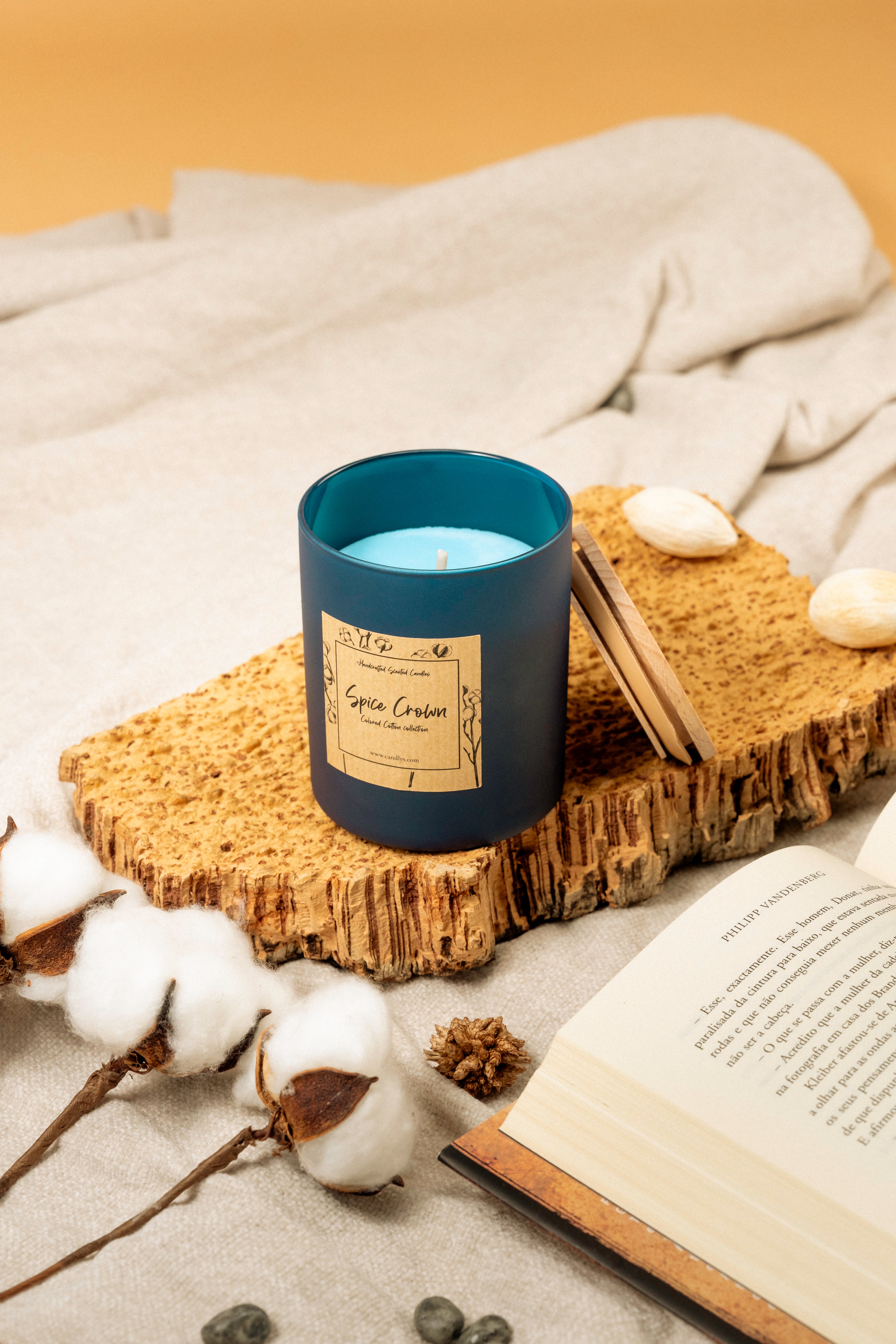 Vela Spice Crown em copo azul-marinho mate, colocada sobre base de cortiça ao lado de um livro aberto e ramos de algodão, criando ambiente acolhedor.