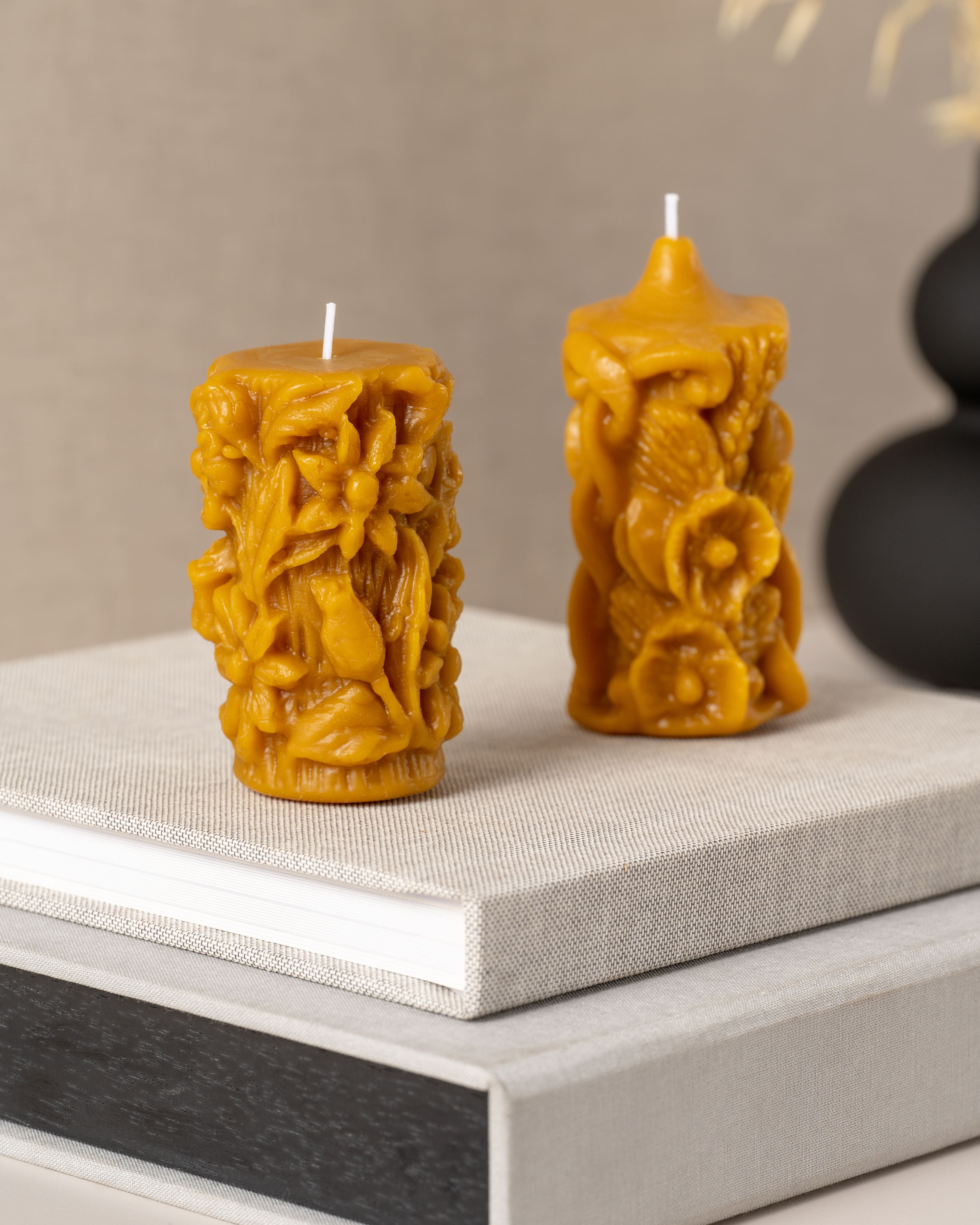 Conjunto de velas artesanais em cera de abelha pura da Candlys. À esquerda, vela altamente detalhada com relevo de flores e folhas do campo; à direita, vela trabalhada com padrões naturais em relevo. Peças únicas que aliam arte decorativa ao aroma naturalmente doce da cera de abelha.