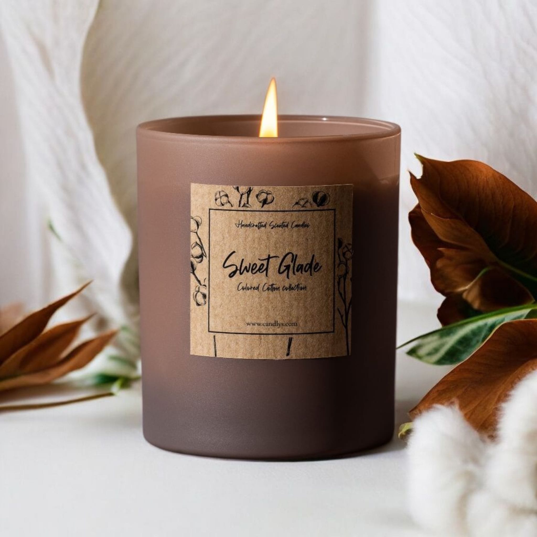 Vela artesanal Candlys Sweet Glade acesa, com cera vegetal e frasco matte, rodeada por algodão e folhas em tons outonais.