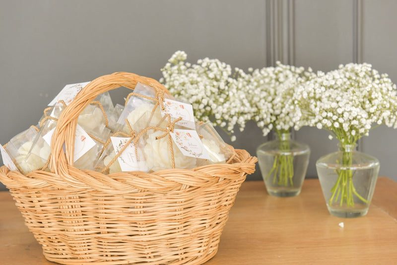 Cesta de verga com velas artesanais Candlys em forma de flor, embaladas individualmente com etiquetas decorativas, ao lado de arranjos de flores brancas em frascos de vidro.
