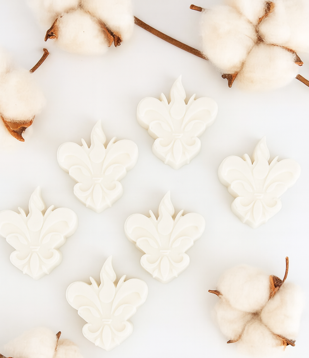 Conjunto de 6 wax melts brancos em forma de flor-de-lis, dispostos num cenário clean com ramos de algodão ao redor. Pertencem à coleção Algodão Colorido — fragrância Bergamota e Jasmim.