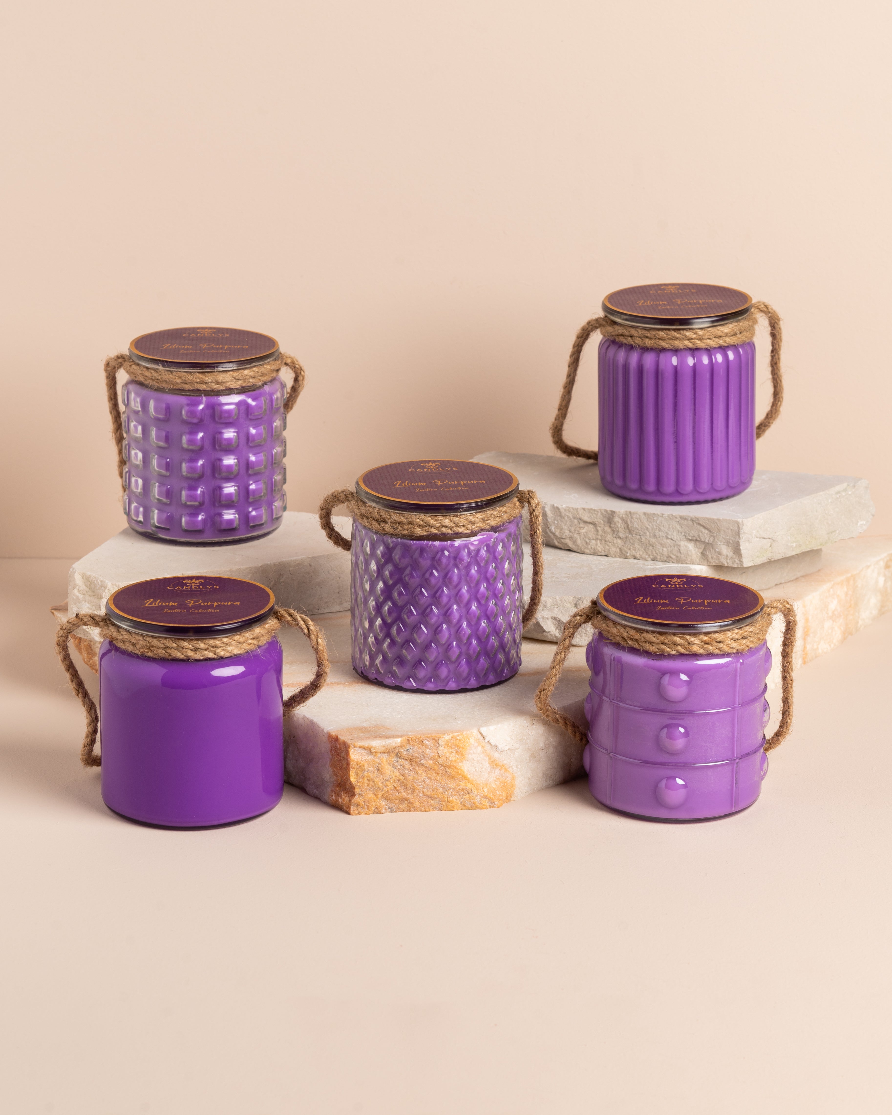Conjunto de quatro velas roxas em frascos de vidro com diferentes relevos e alça de corda, dispostas em base de pedra.