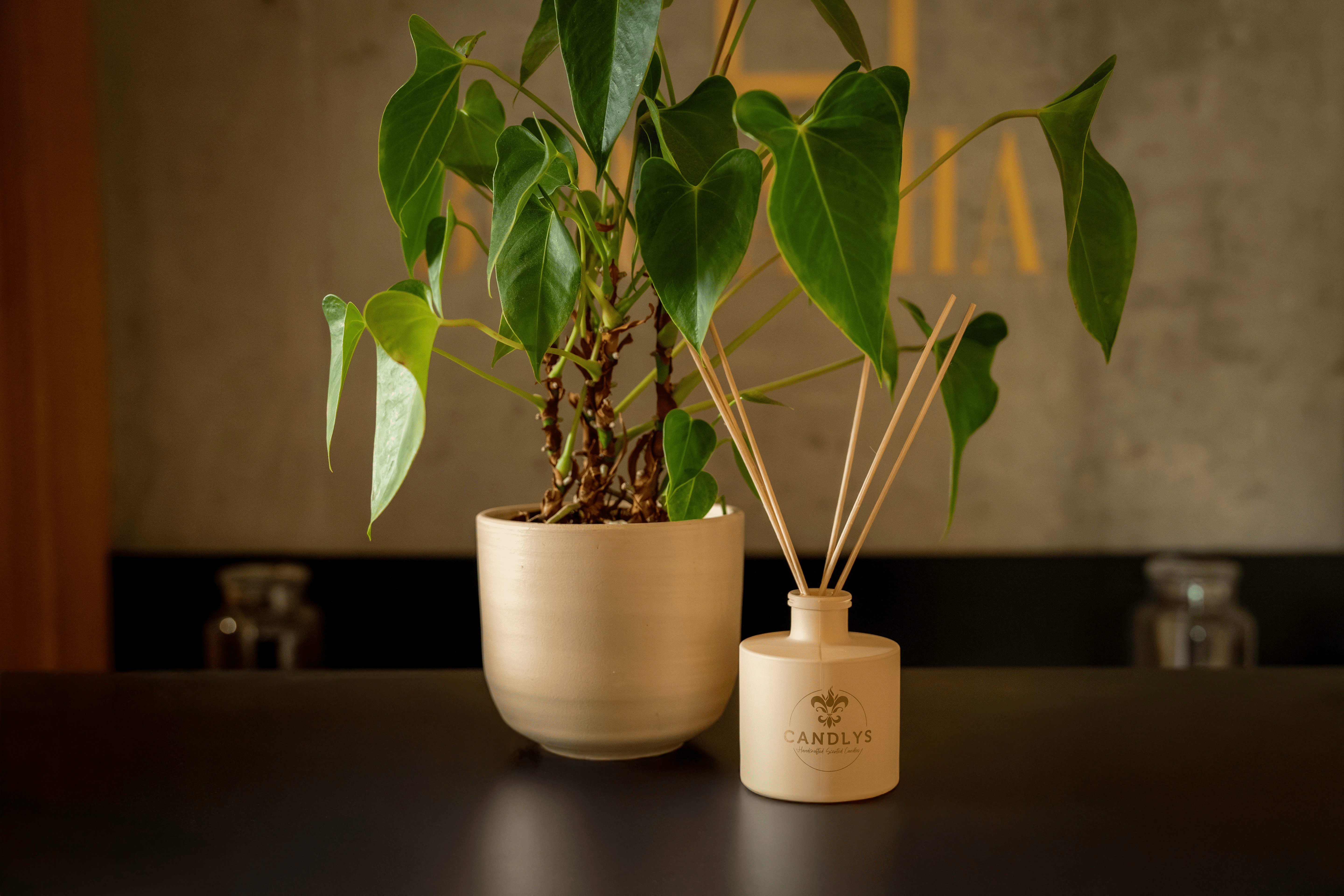 Difusor de aromas Candlys em frasco bege mate com varetas de madeira natural, colocado sobre superfície escura ao lado de um vaso com planta verde, criando um ambiente elegante e fresco.