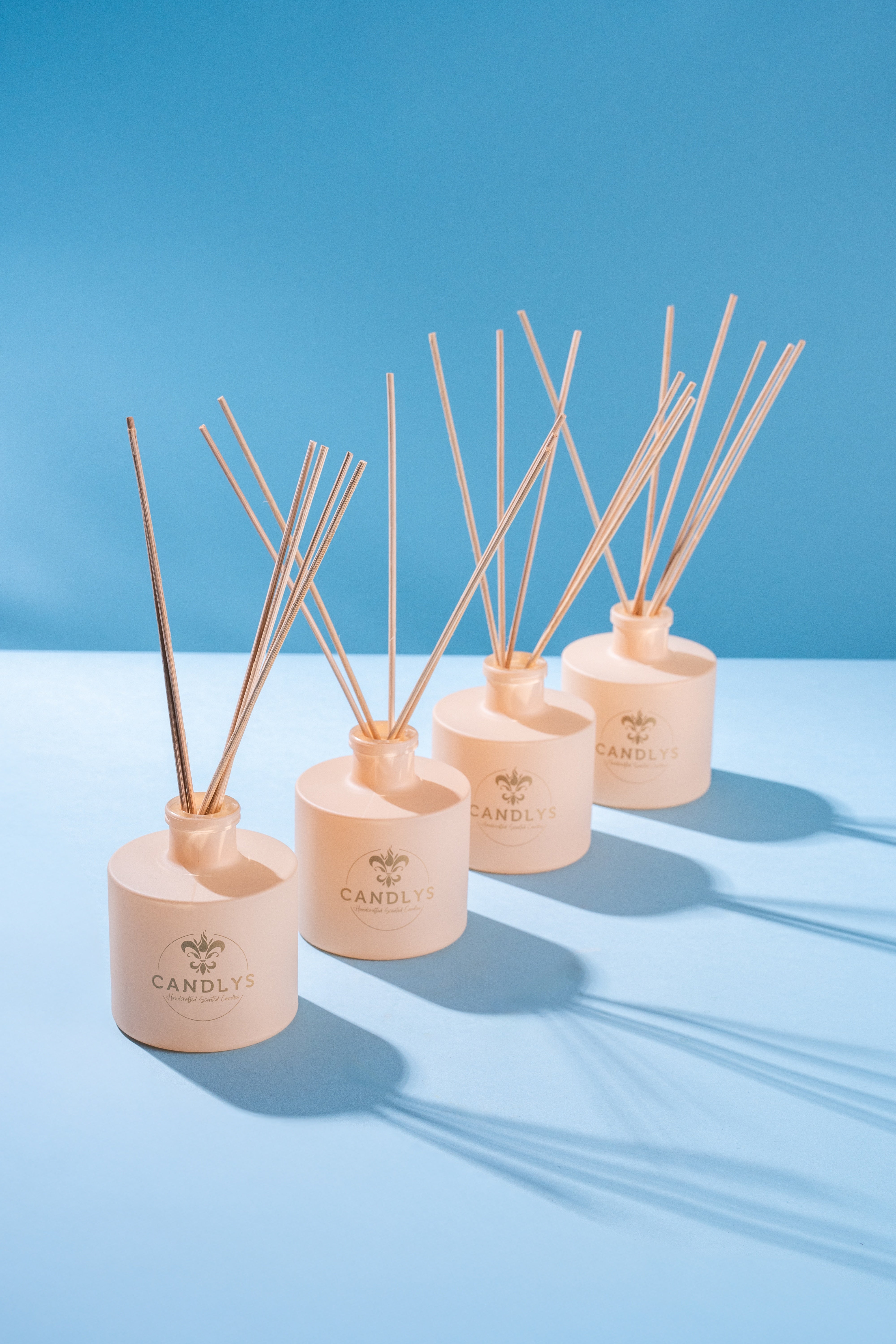Difusores de aromas Candlys em frascos bege mate com varetas de madeira natural, alinhados sobre fundo azul, transmitindo sofisticação e harmonia.
