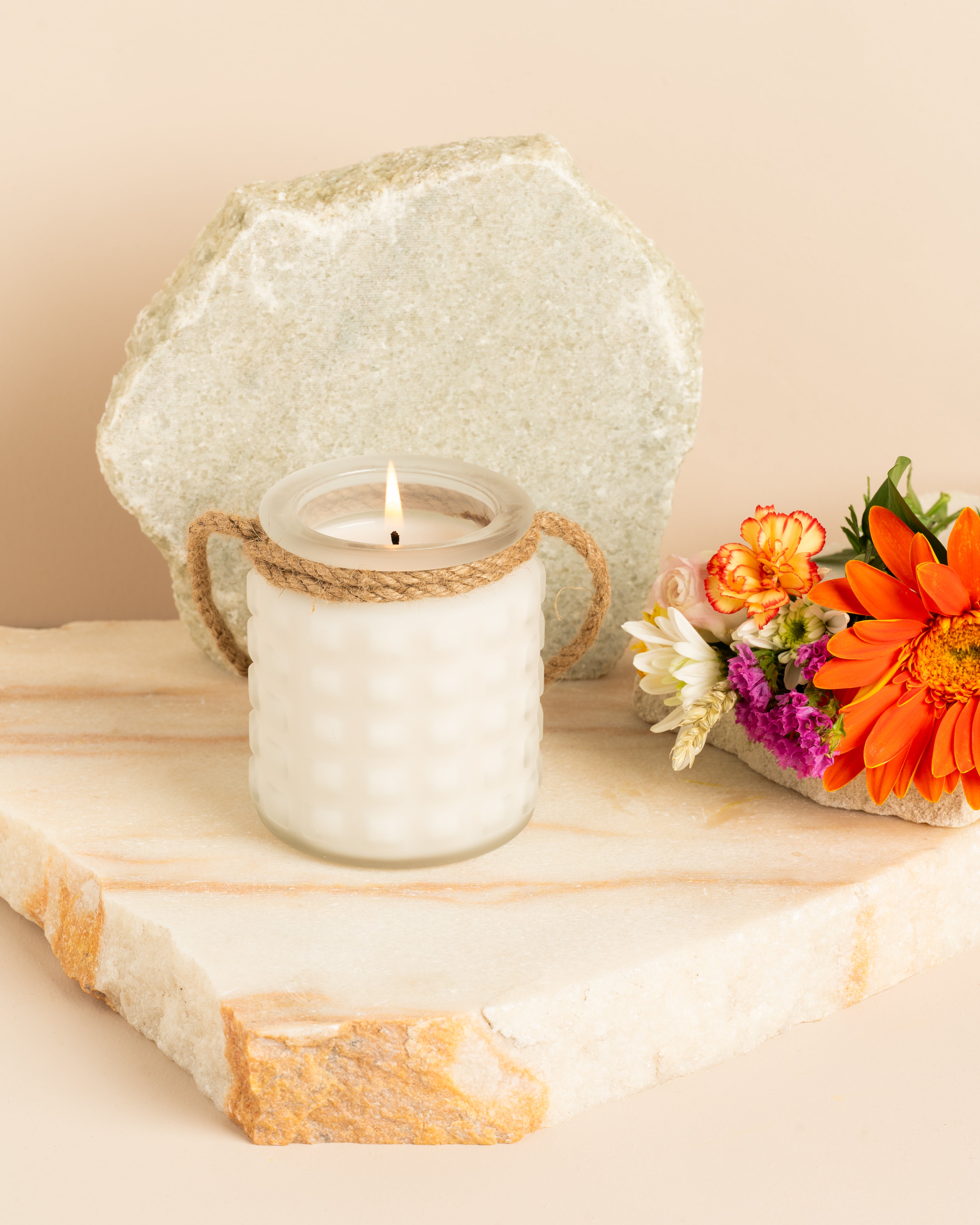 Lanterna de vidro fosco esculpido em forma de pedras preciosas, feita com cera de soja natural branca e fragrância de notas florais campestres e frutadas, decorada com flores coloridas.