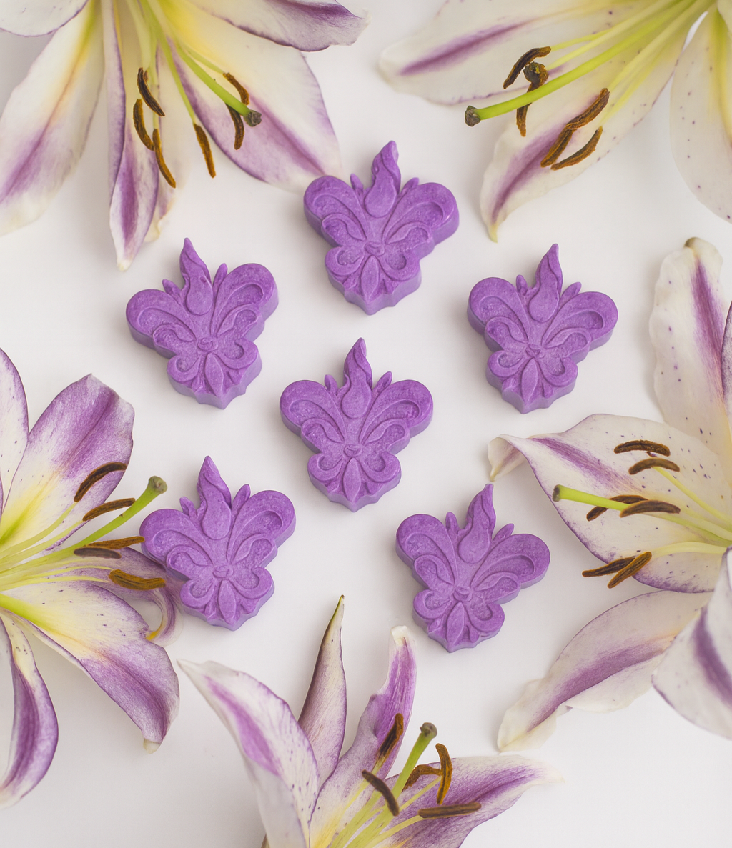 Pack de seis wax melts lilás em forma de flor-de-lis, da coleção Lanterna – Lilium Púrpura, apresentados num cenário clean com lírios lilás dispersos de forma orgânica sobre fundo branco.