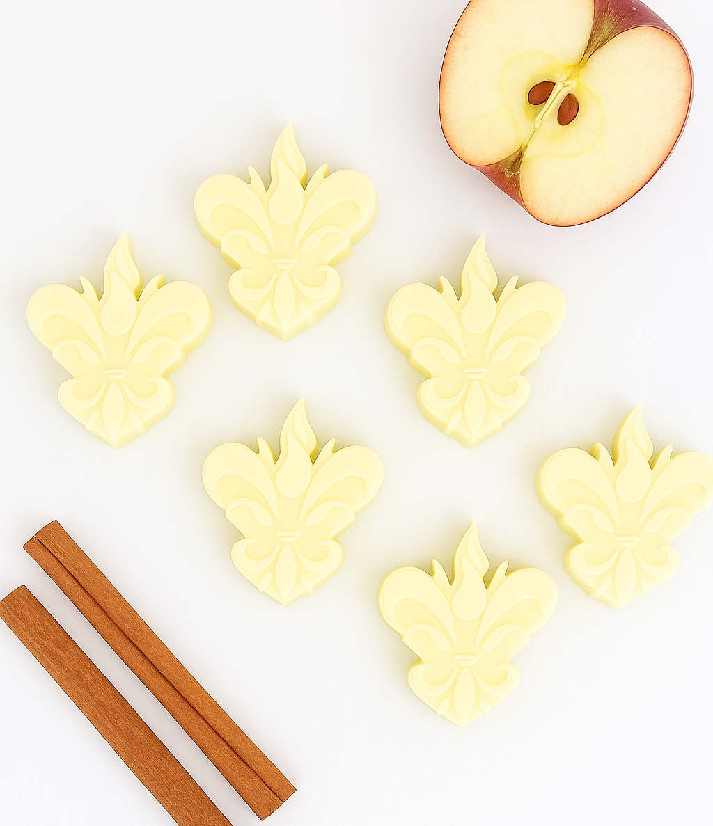 Seis wax melts amarelos em forma de flor-de-lis, dispostos num cenário clean com dois paus de canela e meia maçã vermelha partida — composição minimalista da edição especial Maçã e Canela da Candlys.