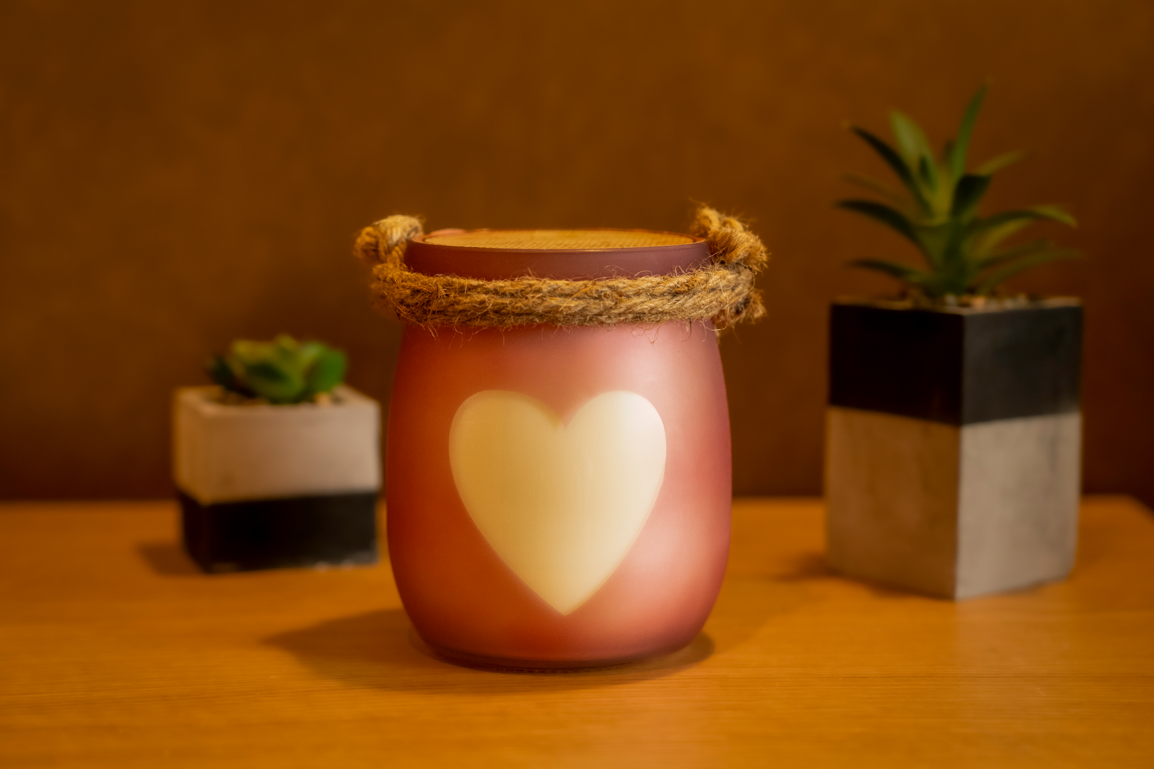 Vela Baunilha & Especiarias em copo cor-de-rosa com coração branco e alça de corda, decorada sobre mesa de madeira ao lado de plantas suculentas.