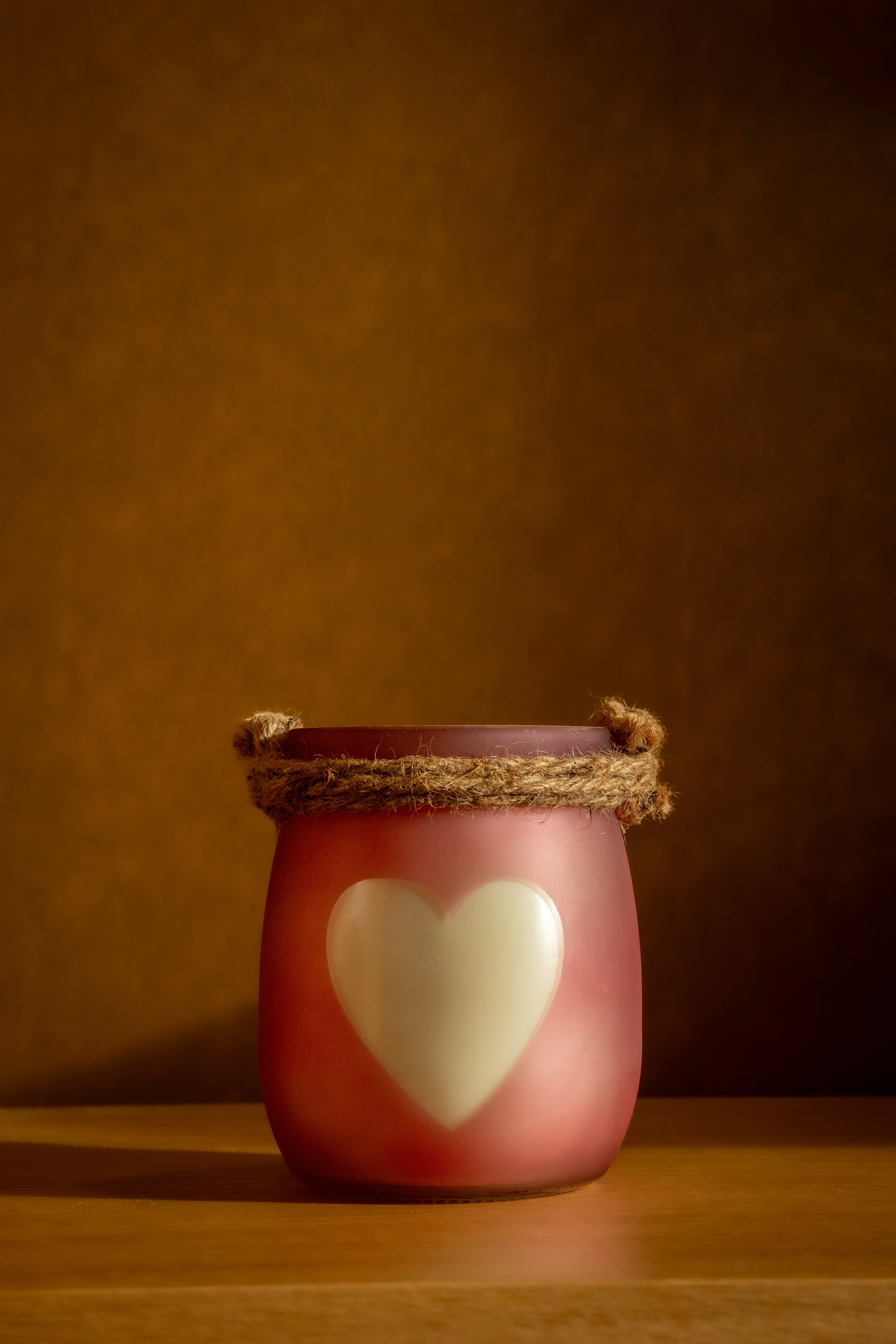Vela Baunilha & Especiarias em copo rosa com coração branco e alça de corda, destacada em cenário quente e acolhedor.
