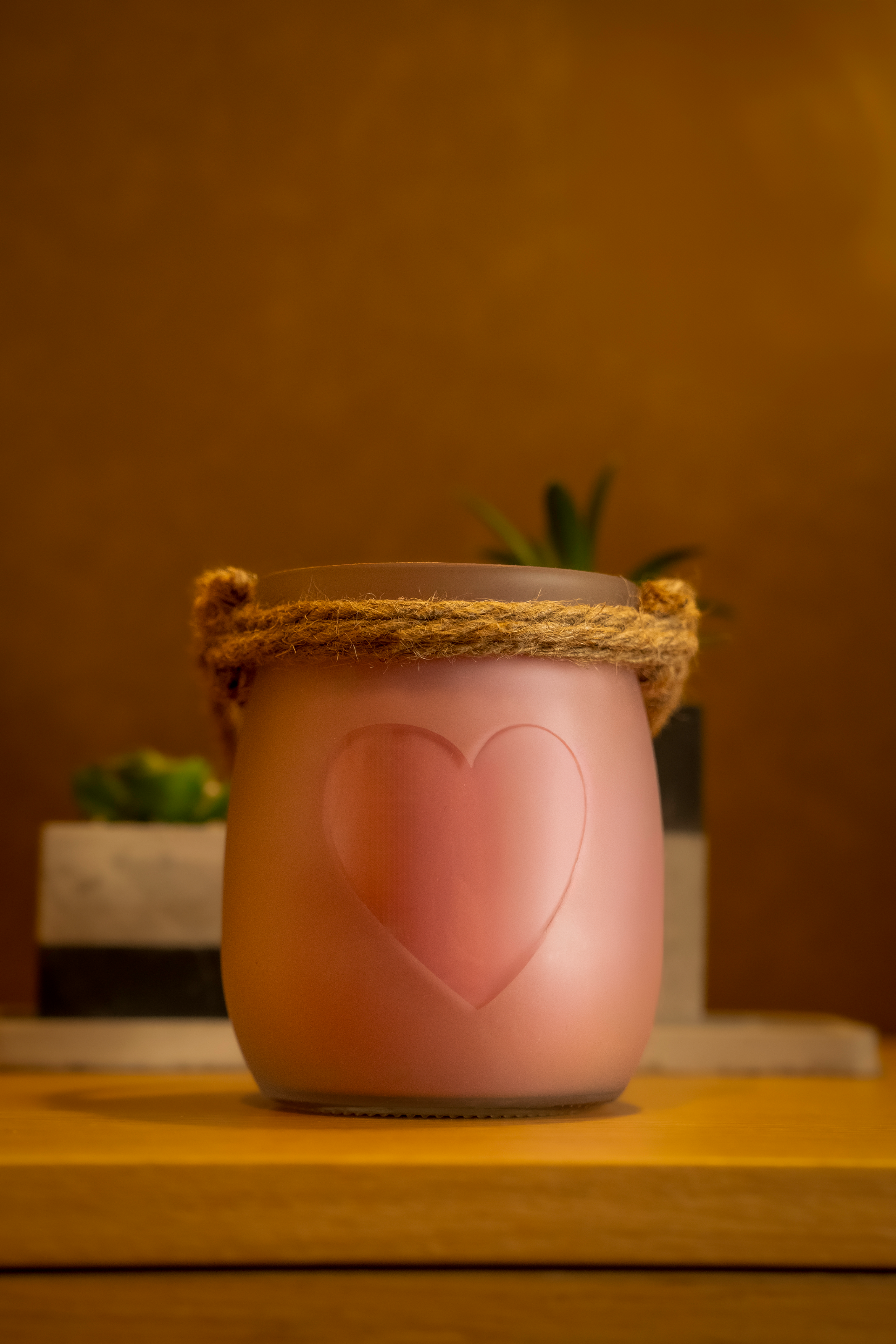 Vela Framboesa & Chantilly em frasco de vidro transparente com cera rosa e coração em relevo, iluminada num cenário acolhedor com fundo quente.