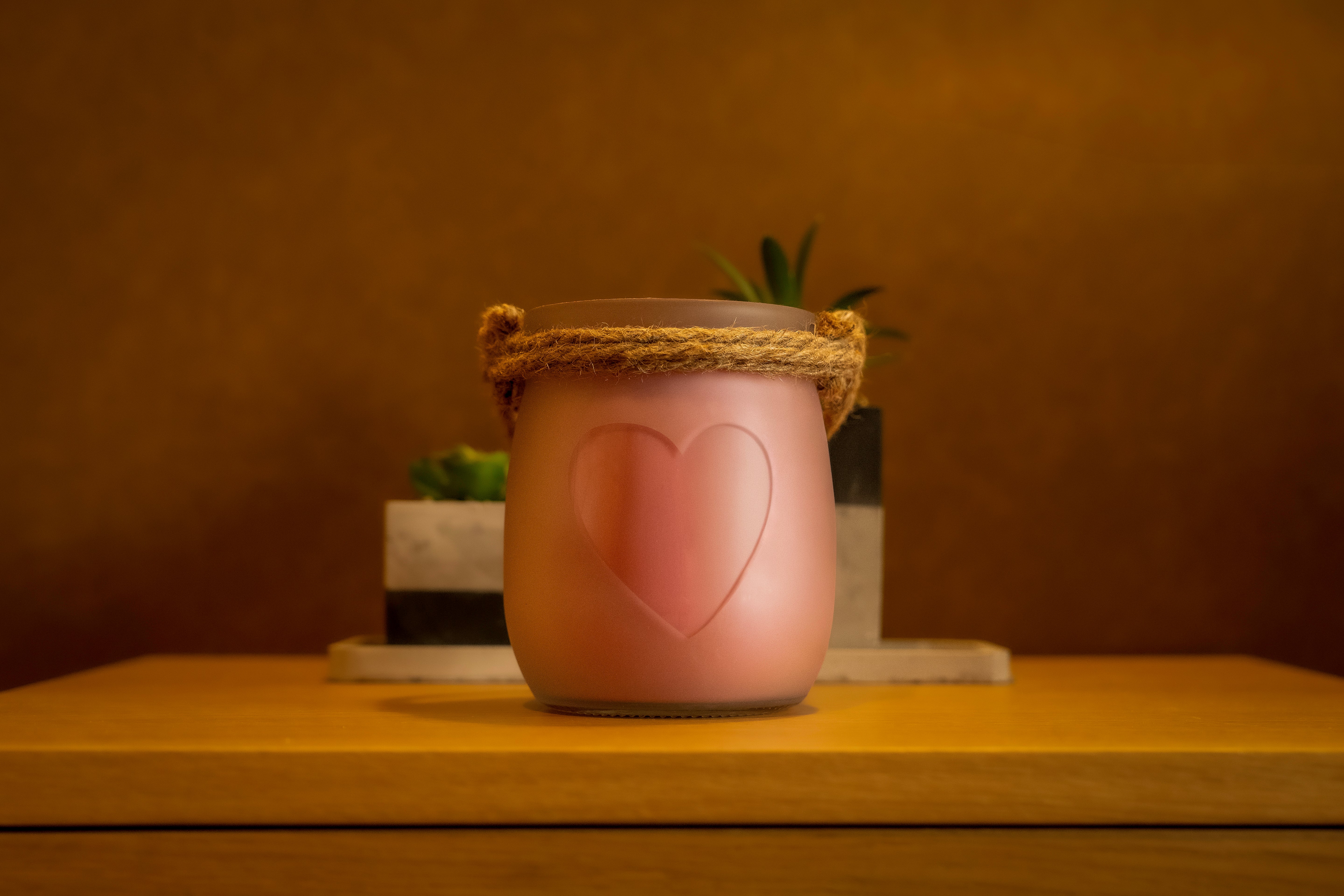 Vela Framboesa & Chantilly em frasco de vidro transparente com cera cor-de-rosa, coração em relevo e alça de corda, decorada sobre mesa de madeira com plantas ao fundo.