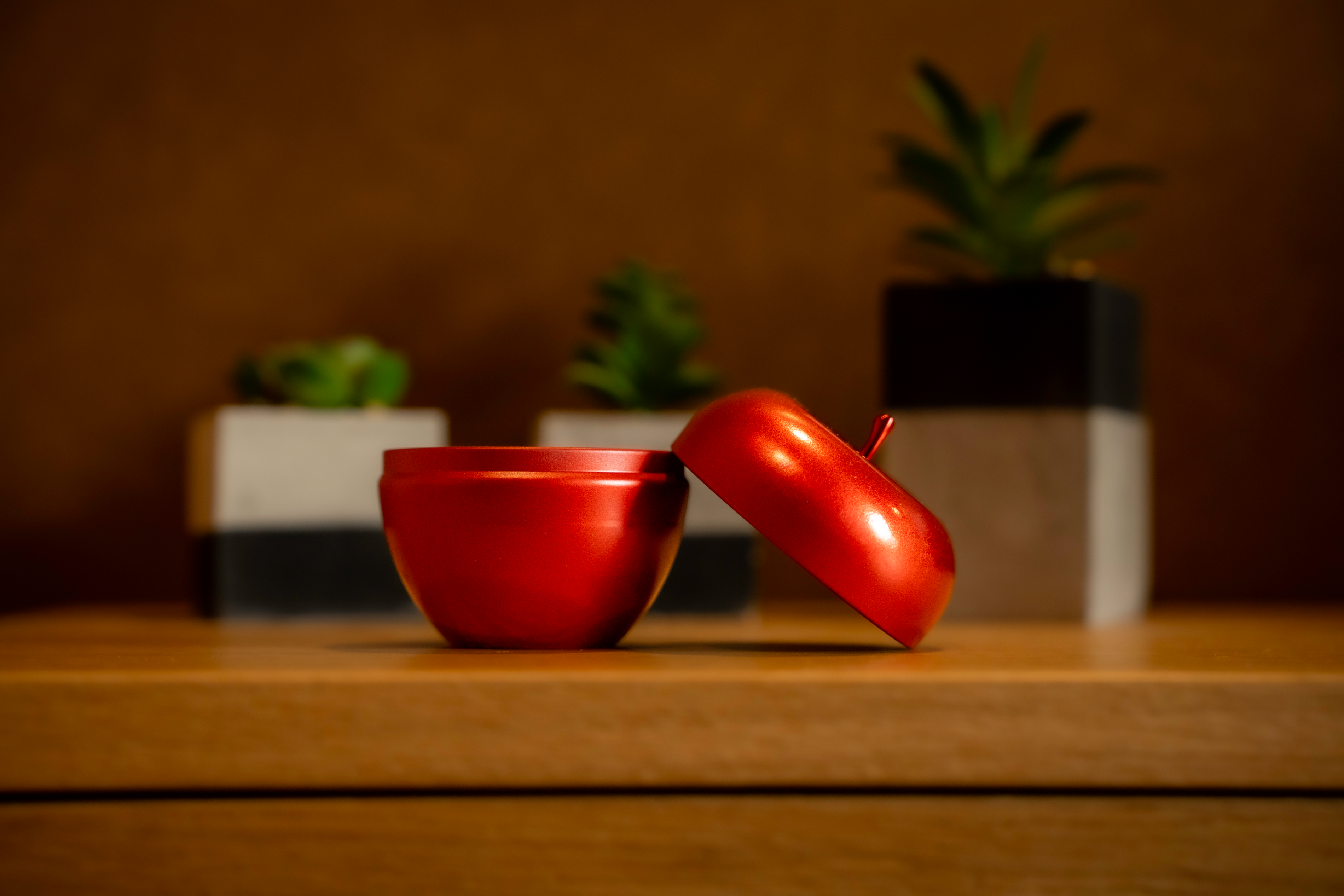 Vela Maçã & Canela em formato de maçã vermelha com tampa aberta, colocada sobre mesa de madeira e fundo quente decorativo.