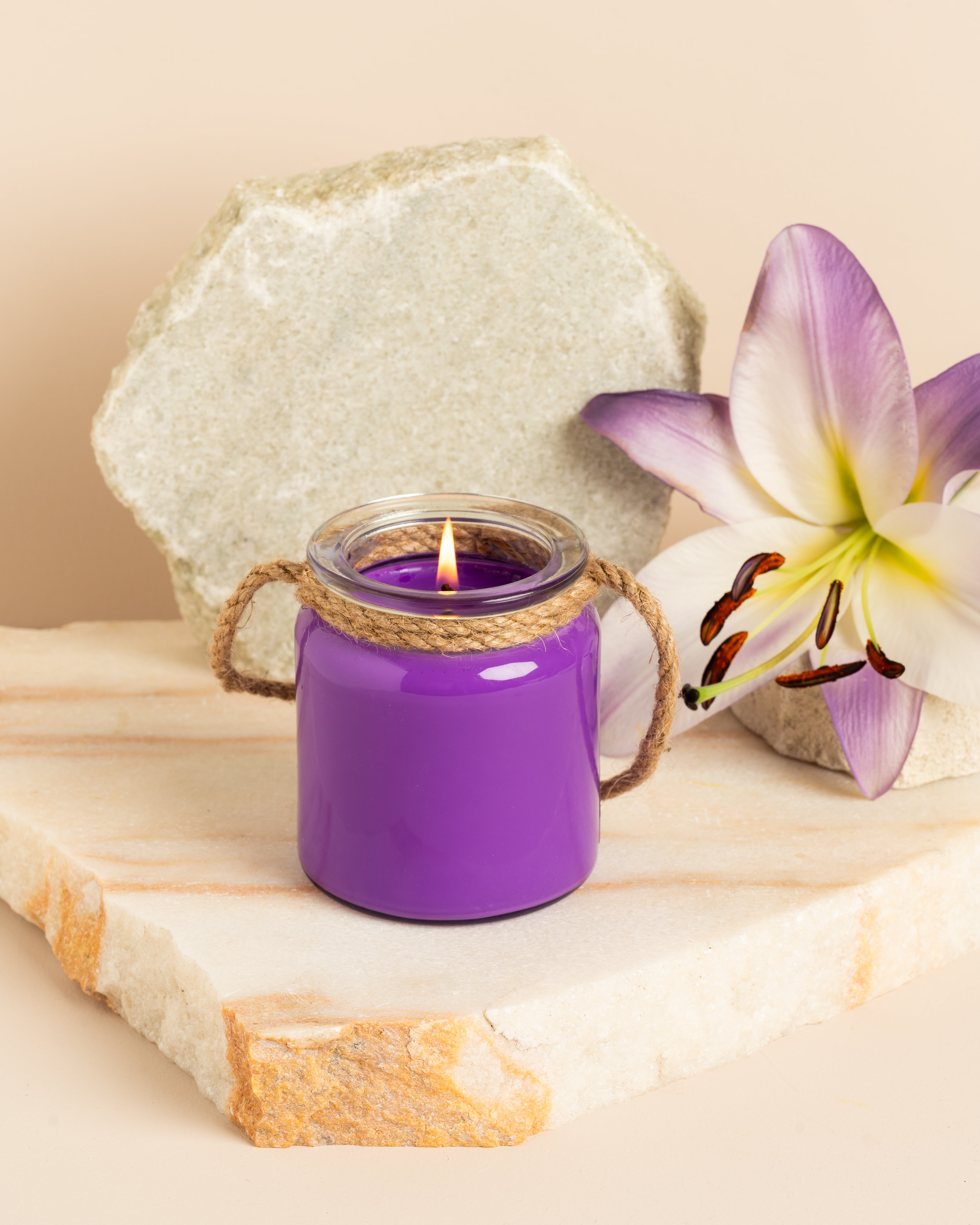 Vela roxa em frasco de vidro com alça de corda, acesa sobre base de pedra, acompanhada por uma flor lilás decorativa.
