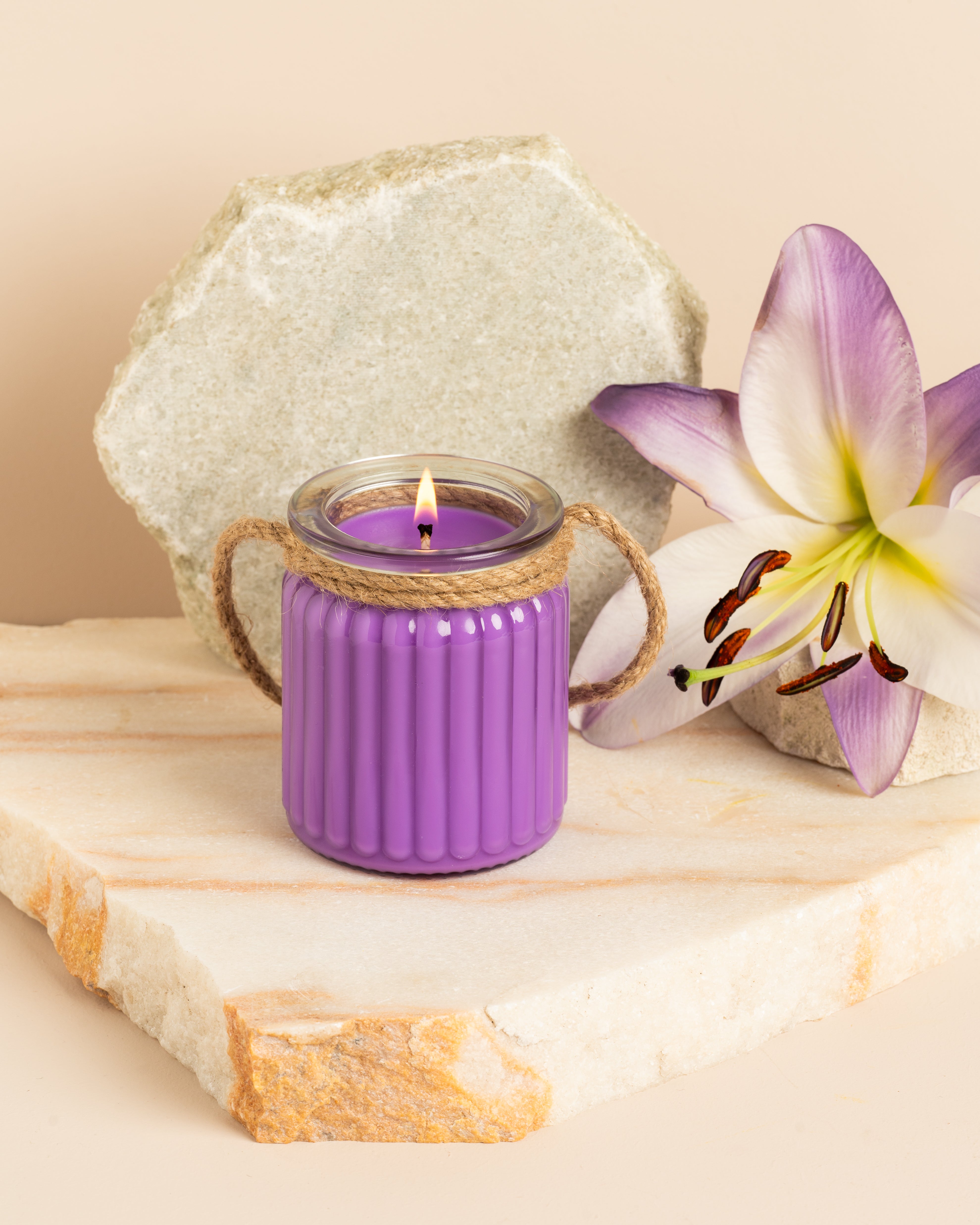 Vela roxa em frasco de vidro ondulado com alça de corda, acesa sobre base de pedra e acompanhada por flor lilás decorativa.