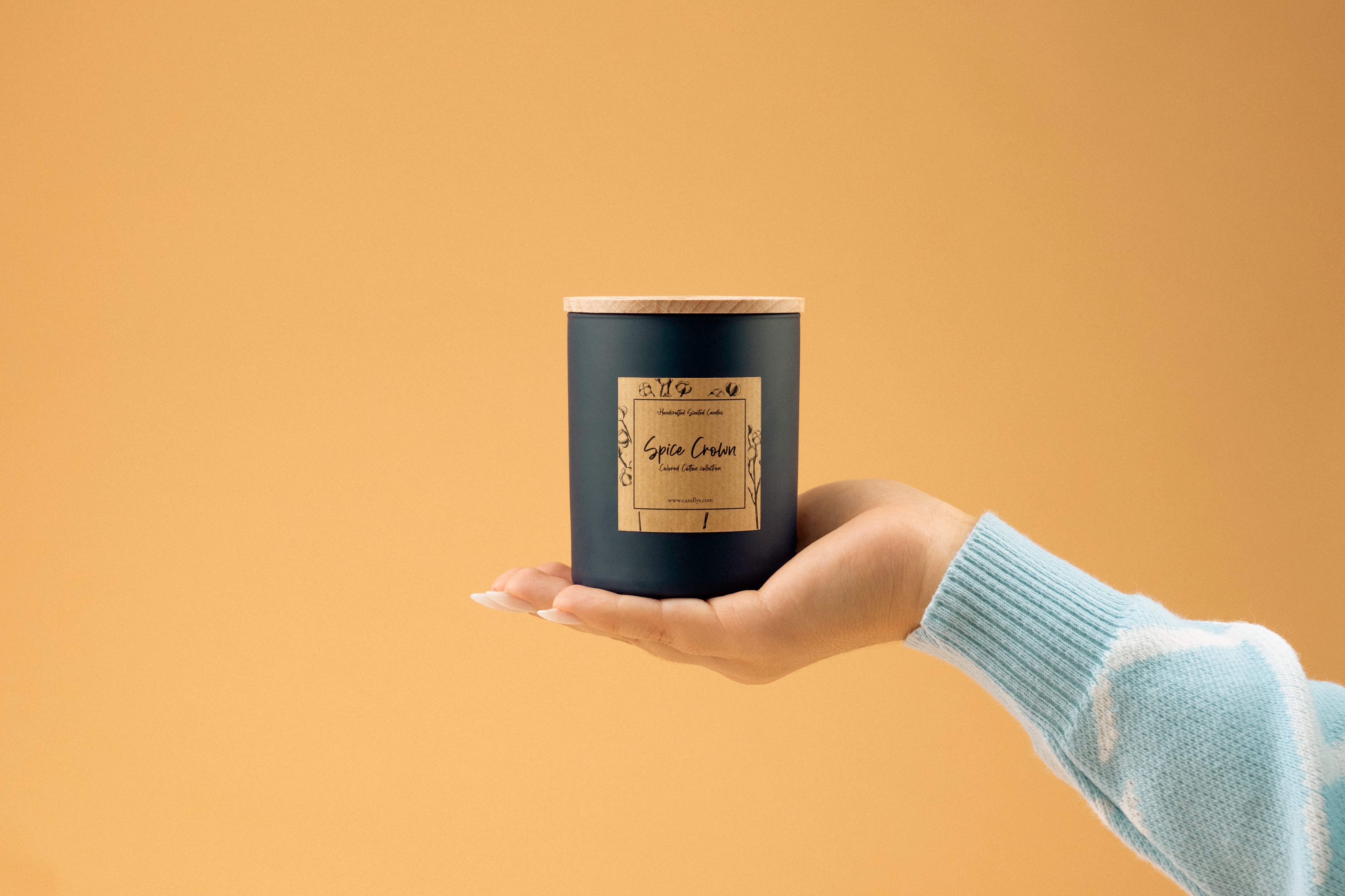 Vela Spice Crown em copo azul-marinho mate, com tampa de madeira e rótulo frontal, apoiada na palma da mão contra fundo bege.