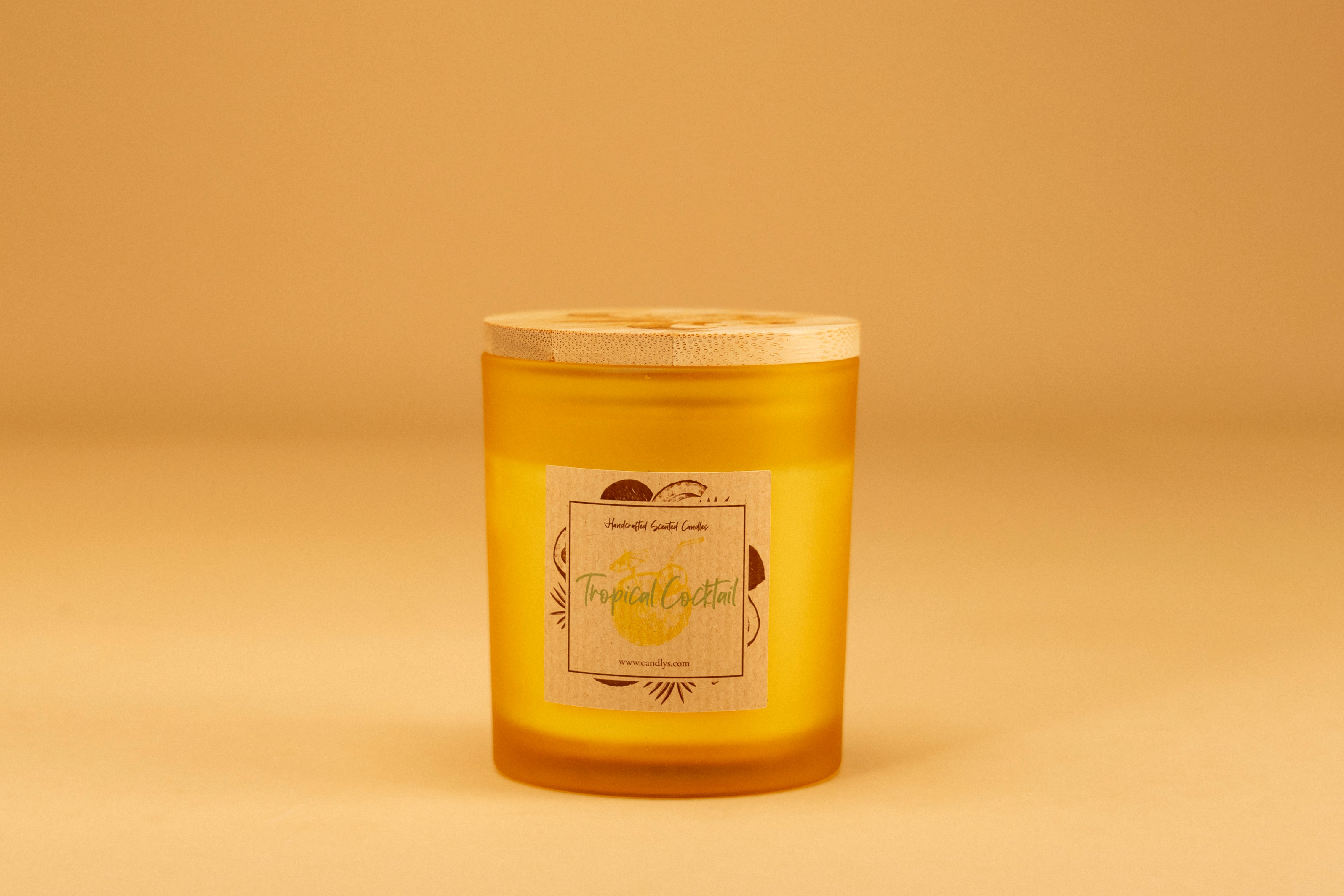 Vela Tropical Cocktail em copo amarelo mate com tampa de madeira e rótulo frontal, fundo bege minimalista.