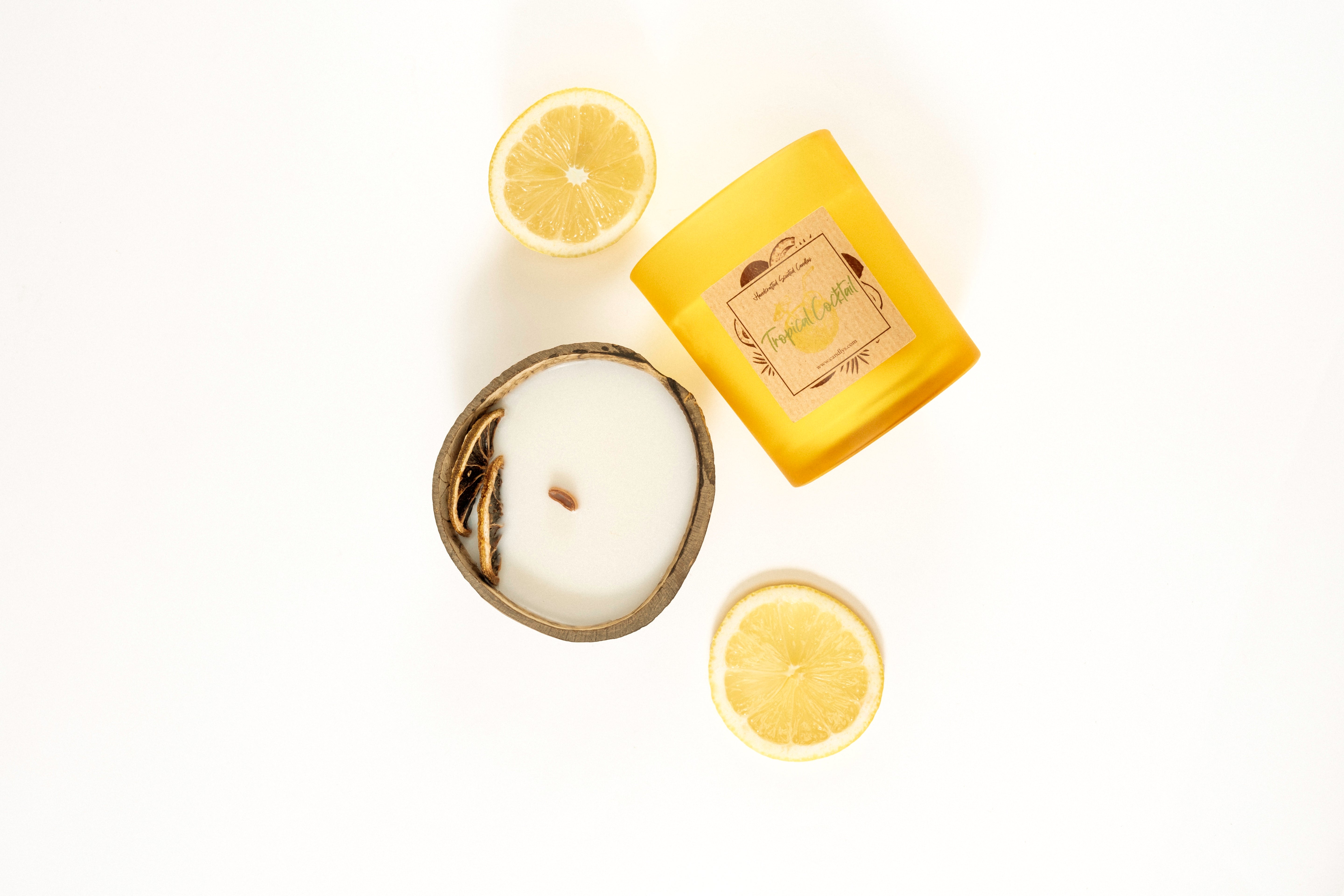 Vela Tropical Cocktail em copo amarelo mate, deitada sobre fundo branco, rodeada por coco e rodelas de limão.