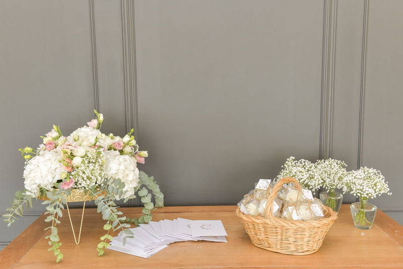 Cesta com velas artesanais Candlys em forma de flor, dispostas sobre uma mesa de madeira com arranjos florais brancos e rosa, ideal para lembranças de eventos.