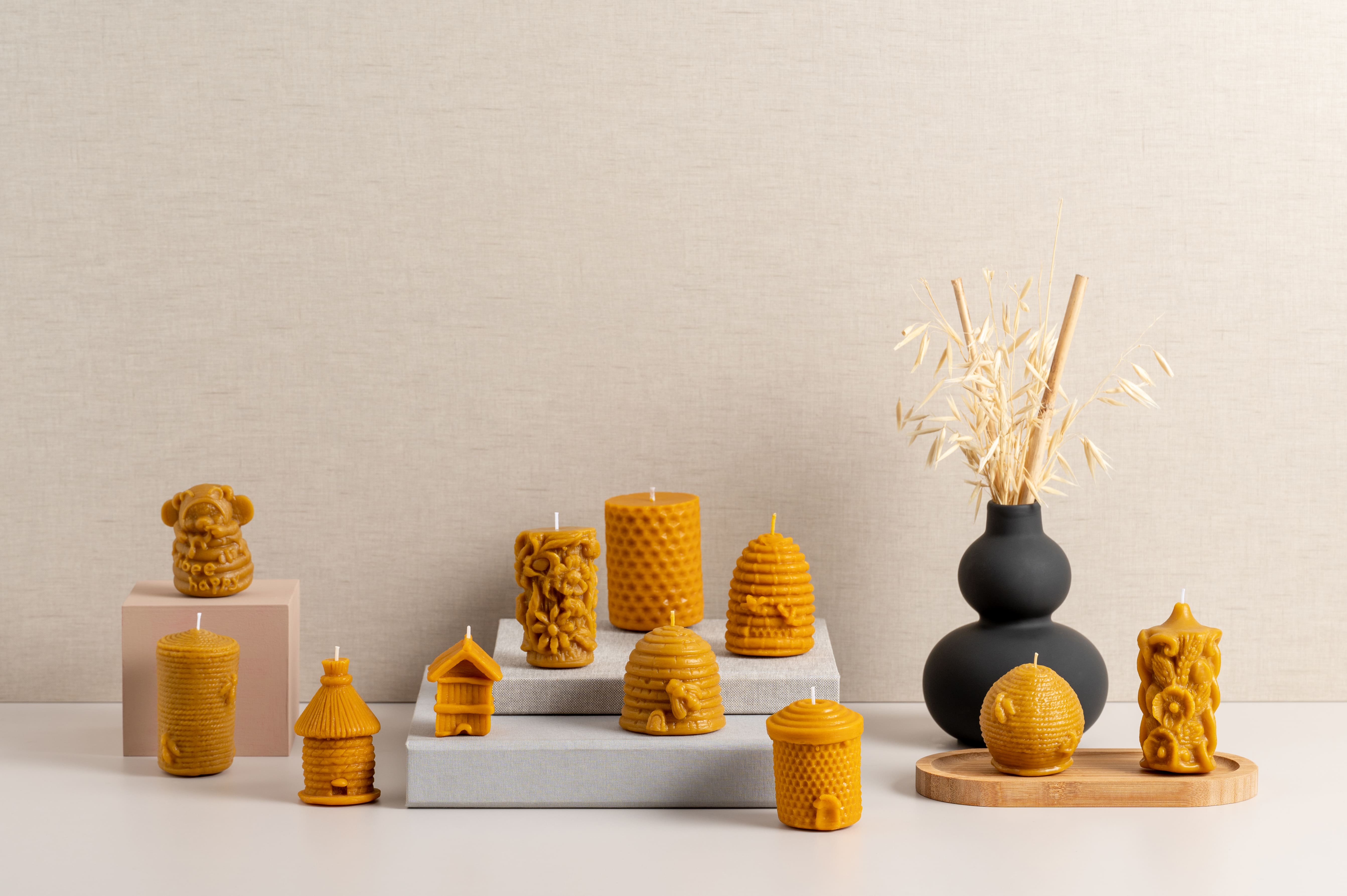 Coleção de velas artesanais em cera de abelha com formas decorativas inspiradas em colmeias, flores, animais e natureza. Expostas sobre blocos neutros e tabuleiro de madeira, com vaso preto e ramos secos ao fundo.